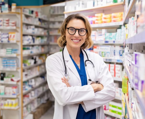 Portrait of mature woman pharmacist at pharmacy wearing labcoat with stethoscope. Happy smiling doctor standing in modern pharmacy drugstore. Friendly young pharmacist owner standing in shop and looking at camera.